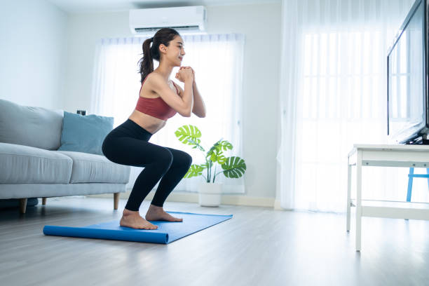 asiatisch schöne junge frau bleibt zu hause und macht aerobic-übungen zu hause. attraktives mädchen, das lockdown-aktivitäten durchführt, durch hocken trainiert, den anweisungen des online-trainers für gesundheit im haus folgt. - hockend stock-fotos und bilder