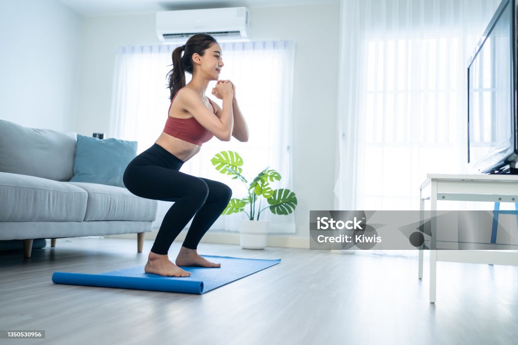 Asian Beautiful young woman stay home, doing aerobic exercise at home. Attractive girl doing lockdown activity, workout by squatting, follow instructions video from online trainer for health in house. Squatting Position Stock Photo