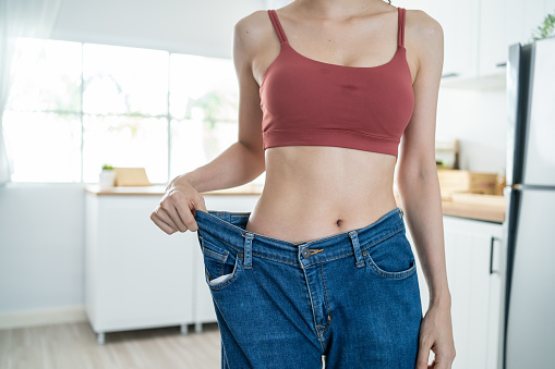 Close up of woman feel happy after lose weight for health in house. Attractive beautiful skinny girl wearing old big jeans with abs abdomen sick packs in kitchen at home. Healthy food and Diet concept