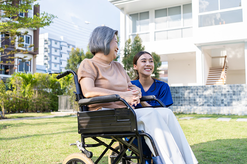 Asian caregiver nurse support old disabled woman on wheelchair outdoor. Beautiful girl doctor help and take care of senior elder mature handicap patient doing physical therapy in park at nursing home.