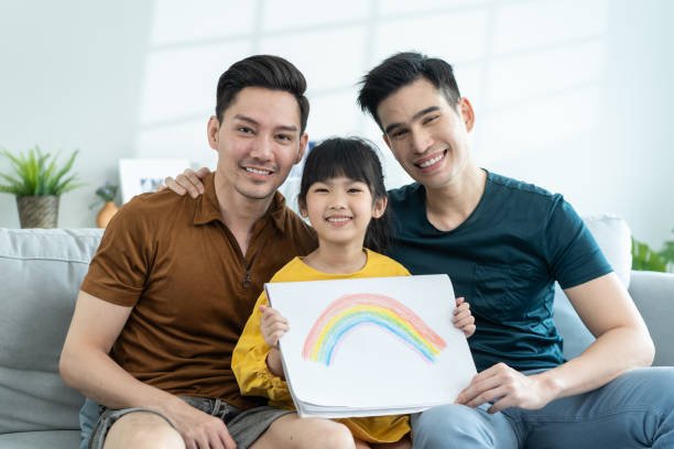 Portrait of handsome man gay family with young daughter in living room. Little adorable girl child sit on sofa with attractive romantic male lgbt couple and shows rainbow drawing picture to two father Portrait of handsome man gay family with young daughter in living room. Little adorable girl child sit on sofa with attractive romantic male lgbt couple and shows rainbow drawing picture to two father gay pride symbol photos stock pictures, royalty-free photos & images