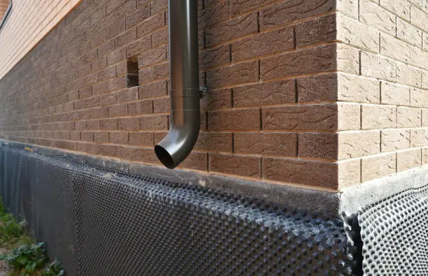 A close-up of a metal downspout, roof gutters downspout installed near unfinished foundation, basement covered with a bitumen waterproofing membrane.