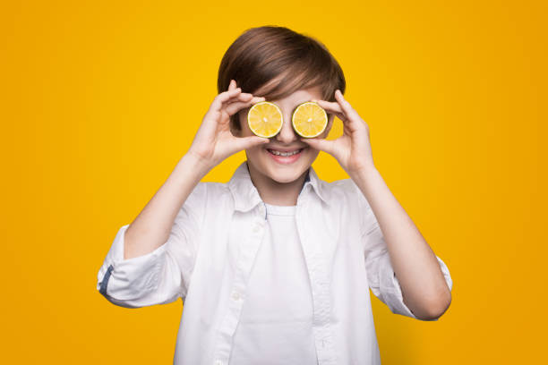 istock Cute boy is covering his eye with a sliced lemons posing on a yellow studio wall smiling at camera 1350570988