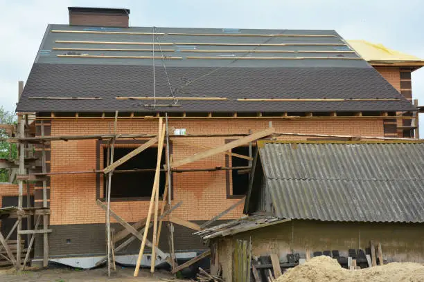 Photo of Asphalt shingles roofing construction. Asphalt shingles installation on the roofing underlayment. Building a new big brick house instead of an old house.