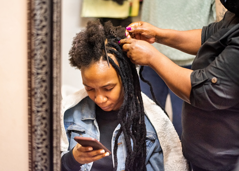 Reflection in mirror of an African woman using mobile phone while getting her hair braided by a hairdresser during a salon appointment