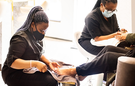 Two beauticians wearing face masks giving pedicure and manicure to a client in beauty salon