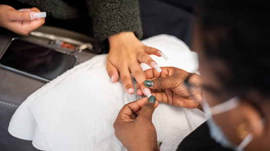 Beautician sticking artificial nails to a female client fingernails for nail extension process in beauty salon