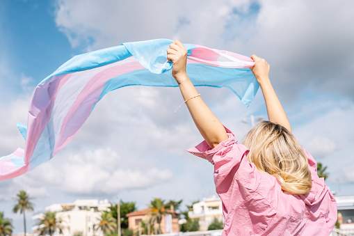 Back view of unrecognizable blond female with transgender flag waving on windy day