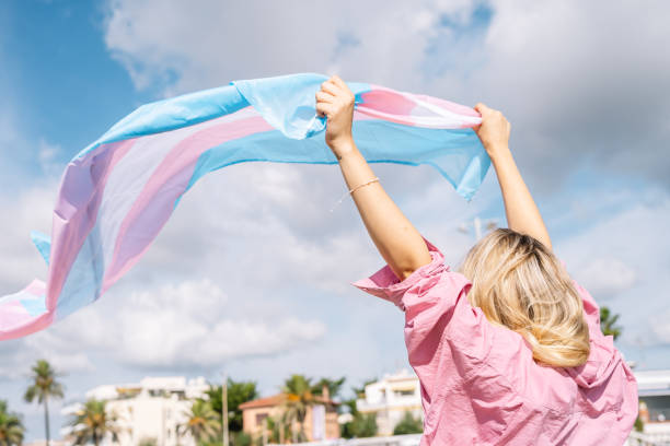 Proud young woman with transgender flag waving on windy day Back view of unrecognizable blond female with transgender flag waving on windy day transgender stock pictures, royalty-free photos & images