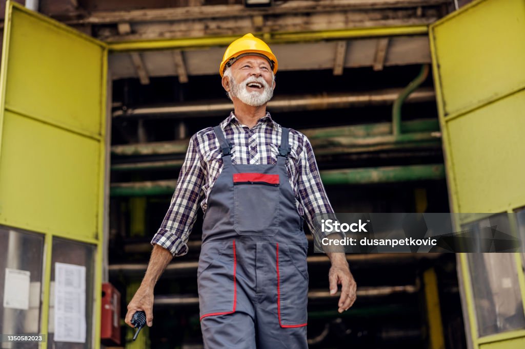 A happy senior factory worker exiting the factory and holding a walkie-talkie in his hands. A factory worker at the factory. After Work Stock Photo