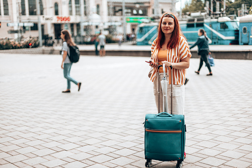 Young caucasian girl in stylish clothes with a smartphone and carry-on travel luggage stands at the station waiting for the train