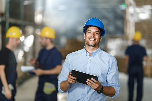 Happy warehouse manager using touchpad in a storage room and looking at camera. There are people in the background.