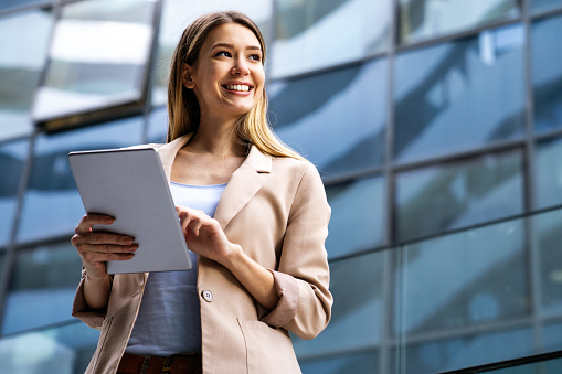 Successful attractive businesswoman using a digital tablet while standing in front of business building. Business people work concept