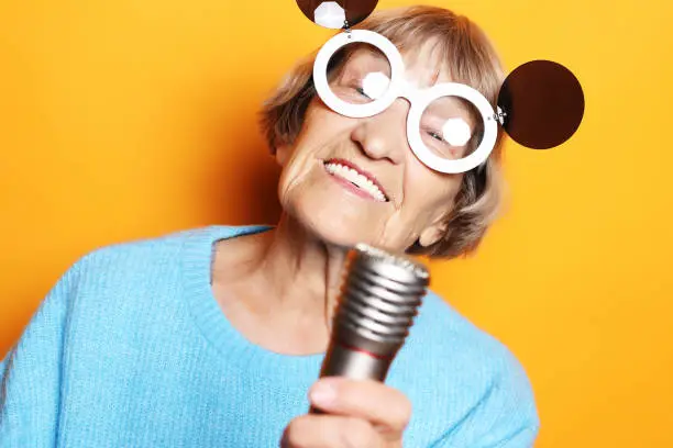 Photo of happy old woman with big eyeglasses holding a microphone and singing isolated on yellow background
