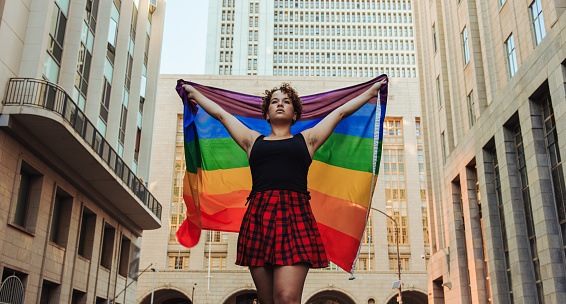 Lesbian woman flying the rainbow flag. Confident young queer woman celebrating gay pride in the city. Young member of the LGBTQ+ community embracing her identity.