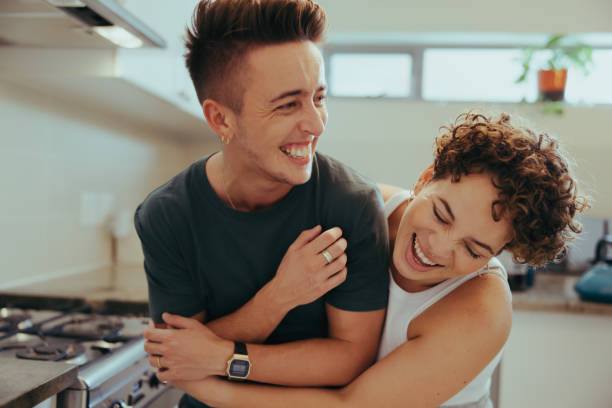 Young queer couple laughing cheerfully in their kitchen Young queer couple laughing together indoors. Happy young queer couple having fun together while standing in their kitchen. Romantic young LGBTQ+ couple bonding fondly at home. transgender stock pictures, royalty-free photos & images