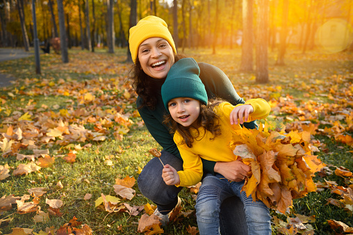 Cheerful woman mother and daughter- adorable baby girl in woolen knitted green yellow hat and sweaters holding a bouquet of falling dry autumnal maple leaves and smiling toothy smile looking at camera