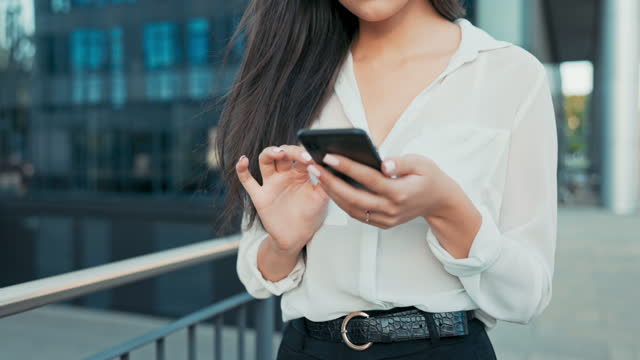Close-up shot of neat hands of elegant woman holding phone, smartphone, woman taps screen, sends message, dressed in shirt walks past glass building of company, business affairs