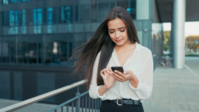Beautiful attractive elegant woman holding a phone in her hands, smartphone, girl taps on the screen, sends a message, wearing a shirt walks past glass building of the company, business affairs