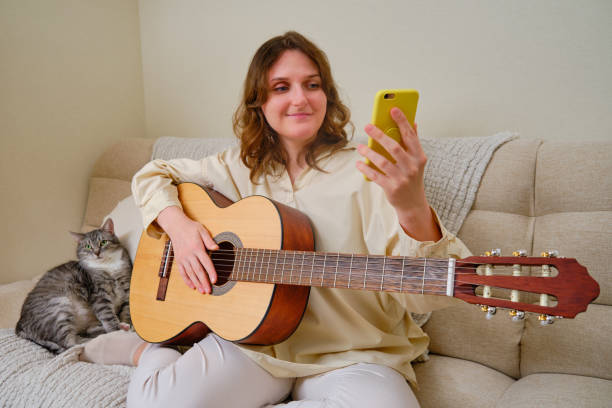 mulher músico com violão olhando para o celular em casa em sofá na sala - mobile phone home video camera video photograph - fotografias e filmes do acervo