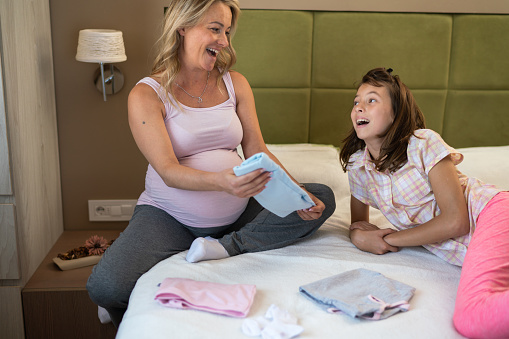 Smiling pregnant mother and her daughter sitting on the bed and preparing baby clothes