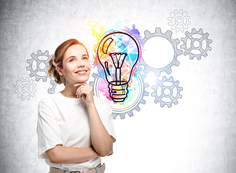 Portrait of smiling European attractive young woman in casual t-shirt pondering about start up and new business ideas. Sketch of colorful light bulb and system of cogwheels.