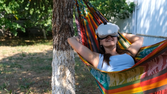 funny woman wearing virtual reality glasses rests in a hammock near a tree, imagining a dream come true, an invented world. The concept of getting experience using VR headset glasses of virtual reality.