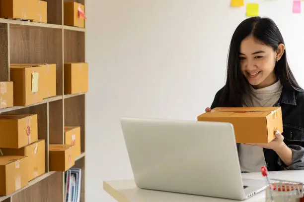 Photo of Young Asian woman holding a work-at-home box and starting a small SME business with online internet. and laptop on the table online shopping and delivery concept
