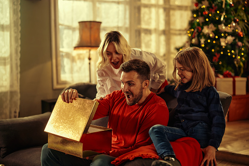 Young adult man opening presents with his wife and son while sitting on sofa during Christmas at home