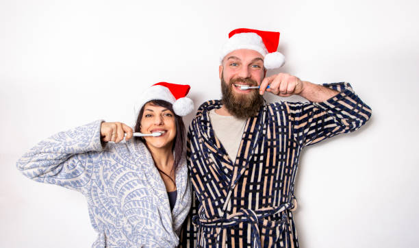 feliz natal! jovem casal adulto comemorando o tempo de natal usando chapéu de papai noel em pé isolado contra a parede de fundo escovando dentes. tempo em família, rostos engraçados e conceito de estilo de vida em feriados - mid adult men portrait hat human face - fotografias e filmes do acervo