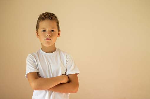 A serious boy of 7-10 years old in a white T-shirt stands with crossed arms on a beige background, copy space.