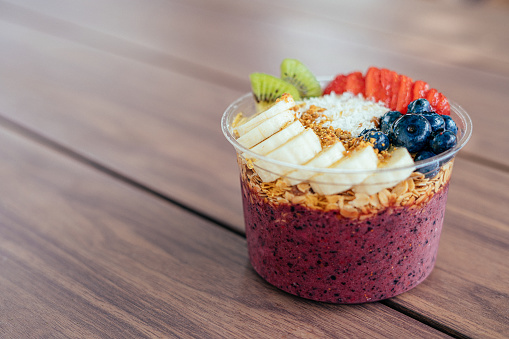 Close-Up Shot of a Healthy Acai Food Bowl with Bananas, Blueberries, Kiwi, Strawberries, Granola, and Bee Pollen on a Wooden Table Outdoors in the Summer with Copy Space and Artistic Bokeh.