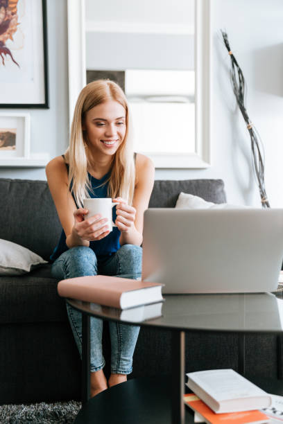 istock Smiling young woman drinking coffee and using laptop on sofa 1350631402