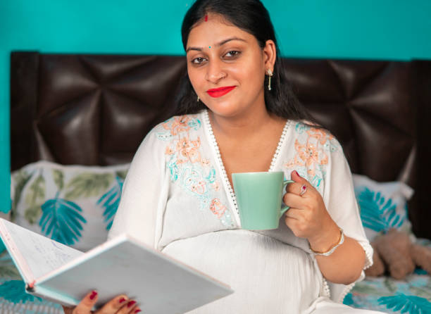 donna incinta felice seduta sul letto a leggere un libro e a prendere un caffè. - baby shower women home interior indoors foto e immagini stock