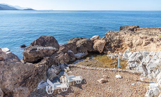 Summer holiday in Greece next to Aegean blue calm sea background. Deserted empty rocky beach with two loungers and an umbrella. White plastic beach chairs for a couple at seaside. Swim, relax.