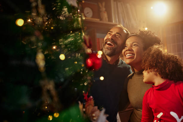 Happy family with cute child decorating Christmas tree together indoors Diverse family decorating Christmas tree. Mixed race girl and black mother helping father decorating the Christmas tree at home christmas excitement stock pictures, royalty-free photos & images