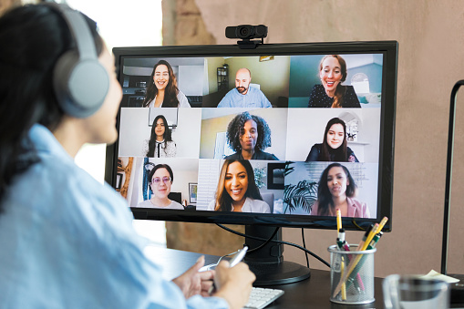 With the focus of the photo on the desktop PC screen, an unrecognizable mid adult woman wears her Bluetooth headphones and attends the virtual meeting with her colleagues.