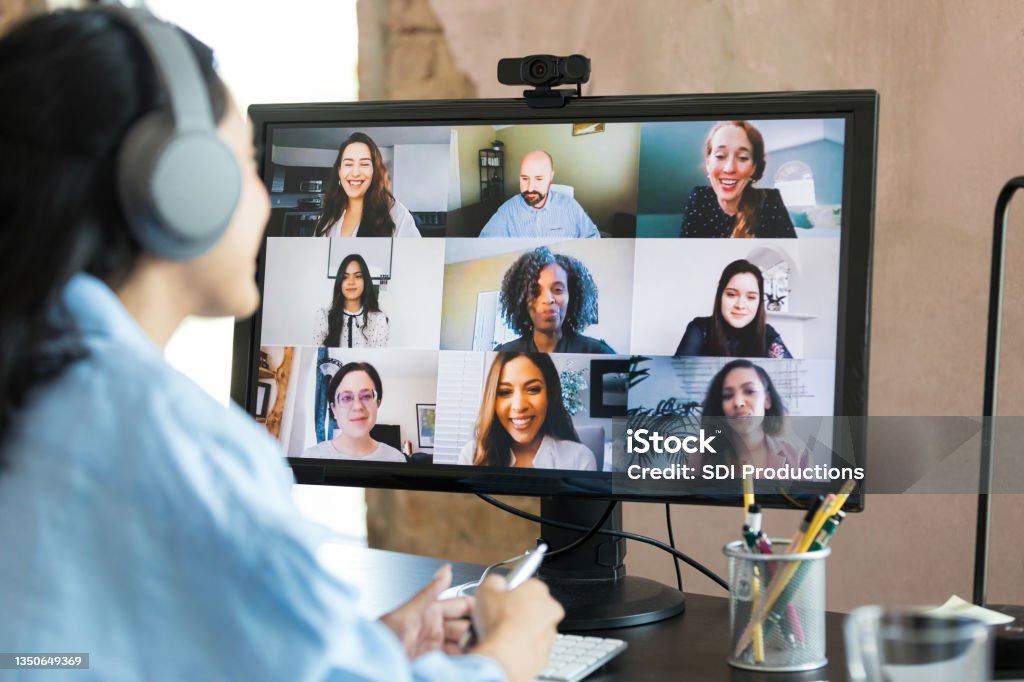Unrecognizable woman wears headphones while attending meeting with co-workers With the focus of the photo on the desktop PC screen, an unrecognizable mid adult woman wears her Bluetooth headphones and attends the virtual meeting with her colleagues. E-Learning Stock Photo