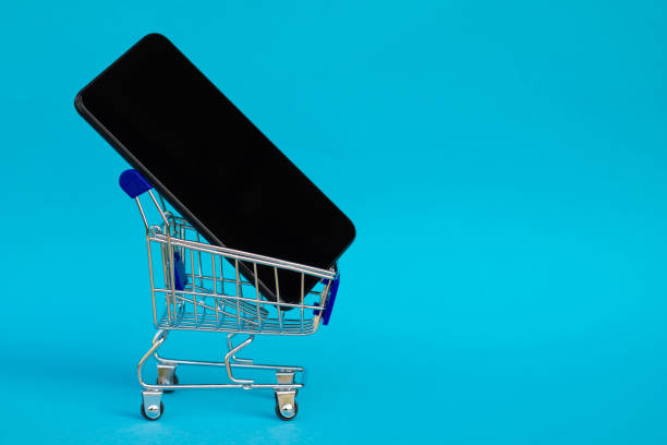 Shopping cart with black mobile phone on blue background. Copy space stock photo