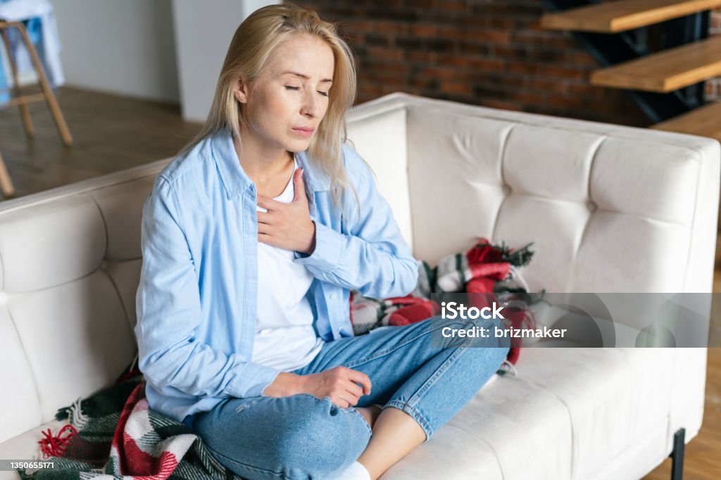 Woman feeling chest pain, sitting at home Exhausted, worried woman feeling pain ache, touching her chest, having heart attack or disease symptom, suffers from heartache sitting at home alone Heart - Internal Organ Stock Photo