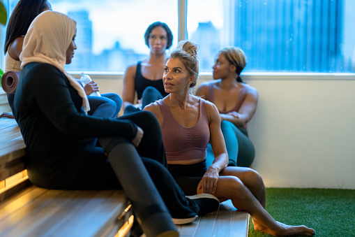 A small group of diverse woman sit scattered on steps after a Yoga class.  They are each dressed in comfortable athletic wear and are talking as they wind down and connect with one another after the class.