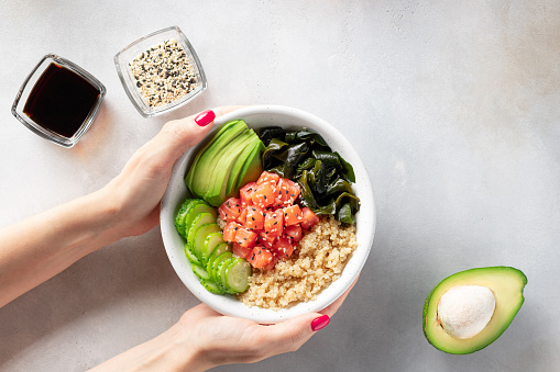female hand holding bowl with quinoa, salmon, avocado, cucumber, wakame seaweed. Trendy healthy asian food. top view, place for text