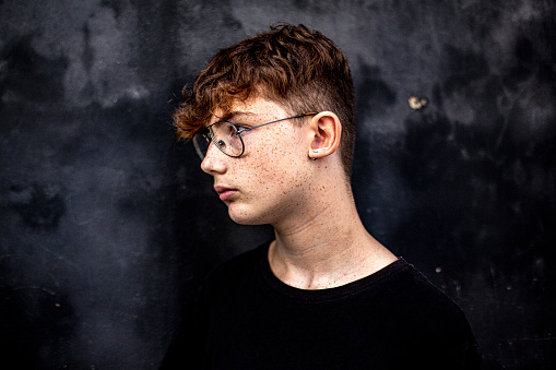 profile of red-haired teenage boy with freckles and eyeglasses against a black wall in the street