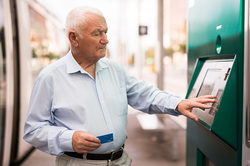 European old man standing on tram stop and using cash machine with credit card.