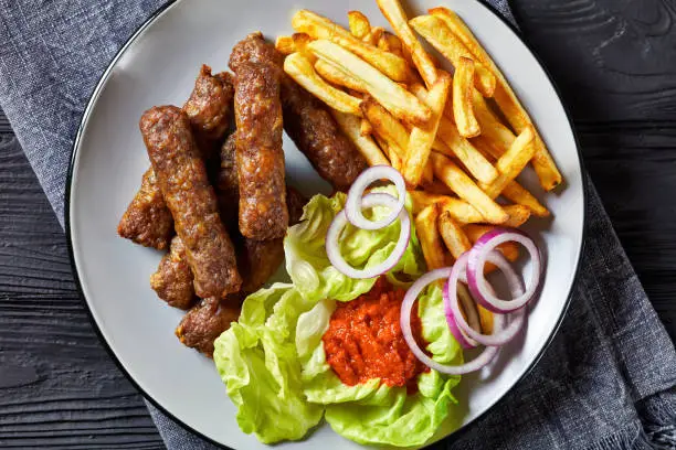 cevapcici, grilled balkan sausages with potato fries, red onion rings, ajvar and fresh lettuce leaves on a plate on a black wooden table, flat lay, close-up