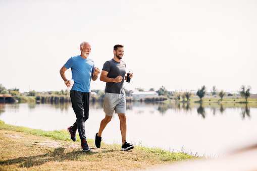 Happy athlete and his senior father jogging by the lake in nature. Copy space.