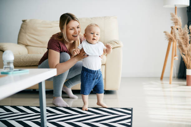Happy mother assisting her small son in taking first steps at home. Happy mother having fun while teaching her baby son to walk at home. Copy space. first steps stock pictures, royalty-free photos & images
