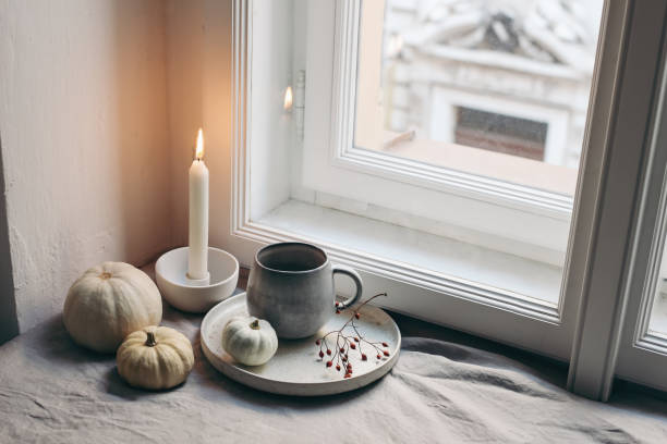 istock Autumn still life. Burning candle, white ceramic candleholder. Moody fall composition. White little pumpkins, cup of coffee and rose hips near window. Scandinavian interior. Halloween, Thanksgiving. 1350692139