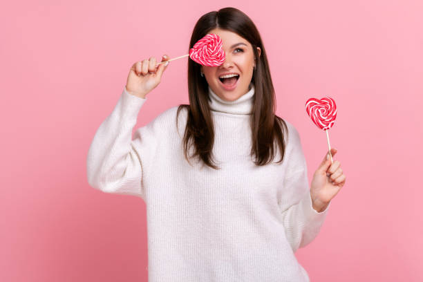 Extremely happy playful brunette woman covering eye with delicious candy in heart shape, looks away. Extremely happy playful brunette woman covering eye with delicious candy in heart shape, looks away, wearing white casual style sweater. Indoor studio shot isolated on pink background. eye candy stock pictures, royalty-free photos & images