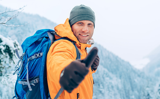Smiling Man dressed bright orange softshell jacket with backpack looking at camera and showing a thumbs-up gesture while he trekking winter mountains route. Active people in the nature concept image.